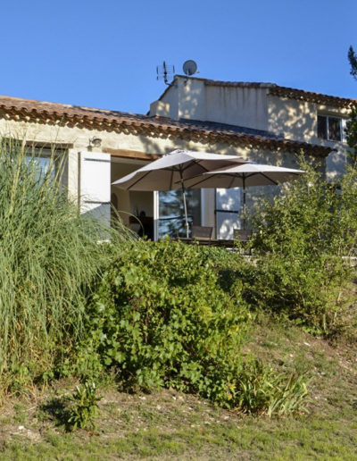 Depuis le jardin végétalisé, vue sur la façade de la pièce de vie avec son revêtement en pierre et ses larges baies vitrées baignant de lumière le salon et permettant un accès direct à l'extérieur de la maison de vacances Les Gavottes située au Revest des Brousses, au cœur de la Provence.