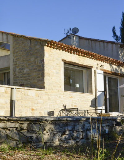 La façade avant de la maison avec son revêtement en pierre lui conférant tout son charme et les larges ouvertures permettant l'entrée de la nature à l'intérieur. Côté entrée, une agréable terrasse avec son barbecue en dur et une double baie vitrée coulissante permet l'accès à la maison par la pièce de vie baignée de lumière pour des vacances agréables au sein de la location Les Gavottes au Revest des Brousses en Provence.