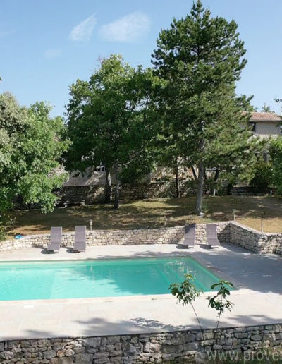 La grande piscine et sa large plage en dalle équipée de bain de soleil dans son écrin de verdure pour des moments détente assurés dans la location de vacances La Poncette située à Reillanne dans le Luberon.