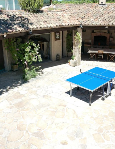 La terrasse ensoleillée avec la table de ping-pong et le auvent couvrant la cuisine d'été avec son barbecue, sa plancha et la grande table pour des raps conviviaux au sein de la location de vacances Le Castellas située à Manosque en Luberon.