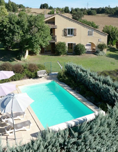 Au premier plan la piscine entourée de ses haies verdoyantes et sa plage en dalle équipée de transat et parasol qui invite à la farniente puis à l'arrière plan la grande maison Les Lavandins comprenant les deux appartements et enfin les champs jusqu'à l'horizon qui compose la location de vacances située à Lurs au cœur de la Provence.