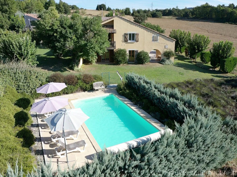 Au premier plan la piscine entourée de ses haies verdoyantes et sa plage en dalle équipée de transat et parasol qui invite à la farniente puis à l'arrière plan la grande maison Les Lavandins comprenant les deux appartements et enfin les champs jusqu'à l'horizon qui compose la location de vacances située à Lurs au cœur de la Provence.