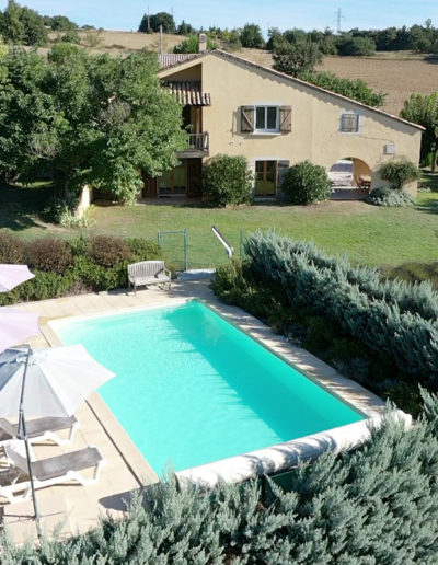 Au premier plan la piscine entourée de ses haies verdoyantes et sa plage en dalle équipée de transat et parasol qui invite à la farniente puis à l'arrière plan la grande maison Les Lavandins comprenant les deux appartements et enfin les champs jusqu'à l'horizon qui compose la location de vacances