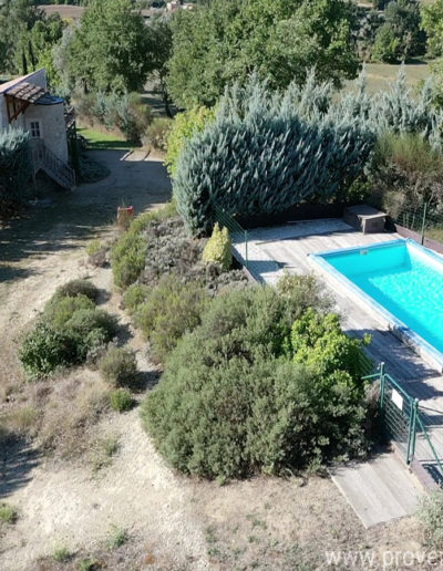 Au premier plan la piscine de La Santoline entourée de végétation avec la maison et sa façade en pierre au second plan et l'environnement arboré au sein de la location de vacances 
