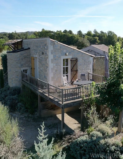 Vue sur la terrasse en bois ensoleillée de la maison La Santoline avec sa façade en pierre et le jardin arboré avec ses essences provençales au cœur du domaine La Tuilière, location de vacances située à Lurs en Provence.