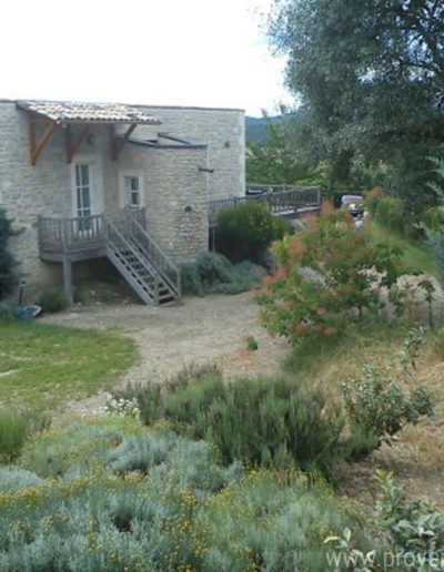 La maison en pierre et sa terrasse en bois qui domine le jardin verdoyant et arboré pour des vacances reposantes dans la location La Santoline située à Lurs en Provence.