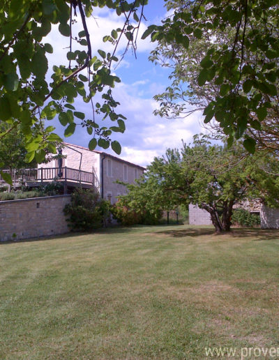 Le jardin du domaine arboré et verdoyant avec la charmante façade en pierre et la terrasse en bois de la location de vacances La Santoline à Lurs, au cœur de la Provence.