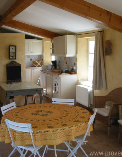 L'espace salle à manger avec sa table ronde recouverte d'une nappe provençale et la cuisine lumineuse à l'arrière plan entièrement équipée de la location de vacances La Santoline située à Lurs en Provence.