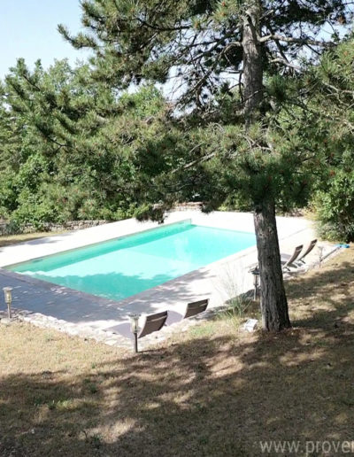 Vue sur la piscine, son eau pure et sa large plage ombragée équipée de bain de soleil au cœur du jardin arboré de la location de vacances La Poncette située à Reillanne dans le Luberon.
