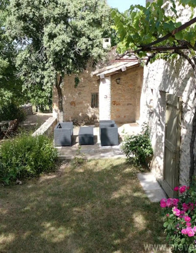 L'espace salon de jardin cosy sur la terrasse en pierre devant la maison et sa façade en pierre agrémentée de végétation et de boutons de roses éclatantes, surplombant l'espace repas sous l'ombre d'un beau chêne en perspectives de bons moments en famille ou entre amis au sein de la location de vacances La Poncette située à Reillanne dans le Luberon.