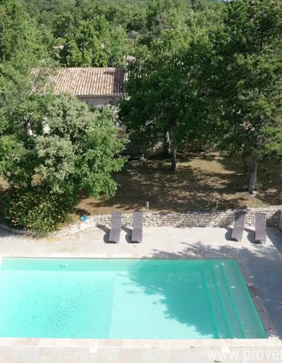 Au premier plan la belle piscine entourée de transats puis à l'arrière plan on devine la maison et le charme de sa façade en pierre à travers la végétation luxuriante du jardin de la location de vacances La Poncette à Reillanne dans le Luberon